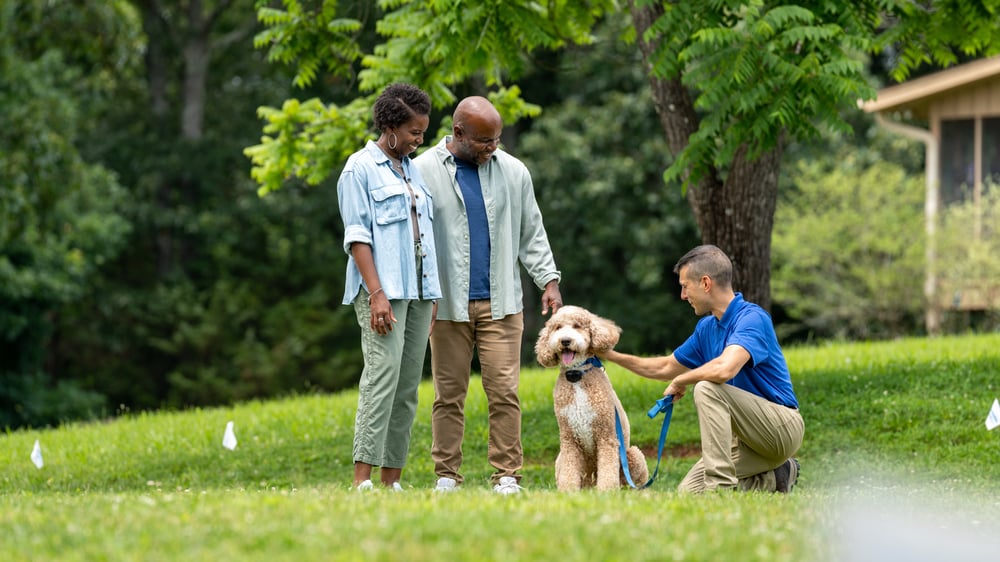 An African American couple are standing in their yard speaking with an Invisible Fence Brand trainer who is kneeling down holding a leash with their cream colored doodle. The doodle is wearing a collar for an Invisible Fence Brand pet fence system. 